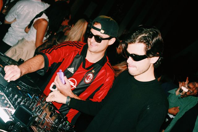 Dos chicos con gafas de sol pinchan hombro con hombro; el de camiseta de fútbol a rayas rojas y negras y gorra hacia atrás sonríe y el de negro mira los platos.