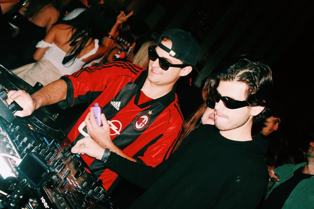 Junto al DJ de camiseta de fútbol a rayas rojas y negras, que sonríe, un chico de negro con gafas de sol mezcla y detrás asoma una chica con gafas oscuras.