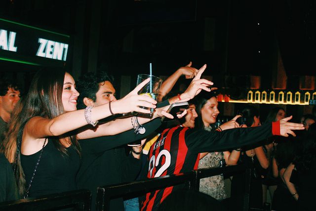 Chicos y chicas estiran los brazos tras la barandilla, entre ellos una chica sonriente con un vaso y un chico con camiseta de fútbol rojinegra, bajo una pantalla de Zentral.