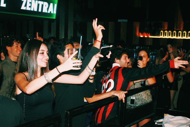 Tras la barandilla, una chica sonríe con un vaso, un chico con camiseta de fútbol rojinegra se gira sonriente y otra chica levanta el móvil, bajo la pantalla de Zentral.
