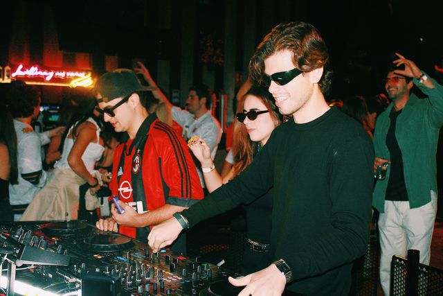Un chico de negro con gafas de sol sonríe mientras mezcla junto al DJ de camiseta de fútbol a rayas rojas y negras; entre ambos, una chica con gafas de sol.