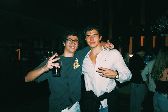 Dos chicos posan abrazados mirando a cámara y hacen la V con los dedos; el de gafas sonríe con un vaso en la mano y el otro lleva camisa blanca.