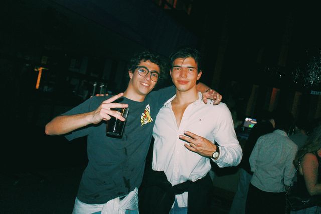 Un chico con gafas y un vaso y otro con camisa blanca y una prenda negra anudada a la cintura posan abrazados mirando a cámara, ambos haciendo la V con los dedos.