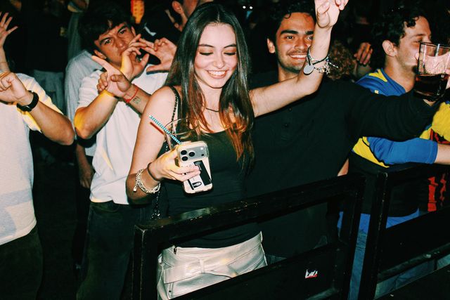 Una chica de top negro sonríe con los ojos cerrados y un brazo en alto, con un vaso y el móvil en la otra mano, rodeada de chicos tras la barandilla.