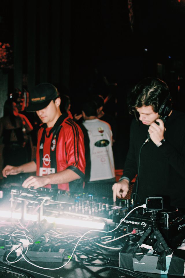 El DJ de camiseta de fútbol a rayas rojas y negras mezcla junto a un chico de jersey negro que se pega un auricular de mano a la oreja.