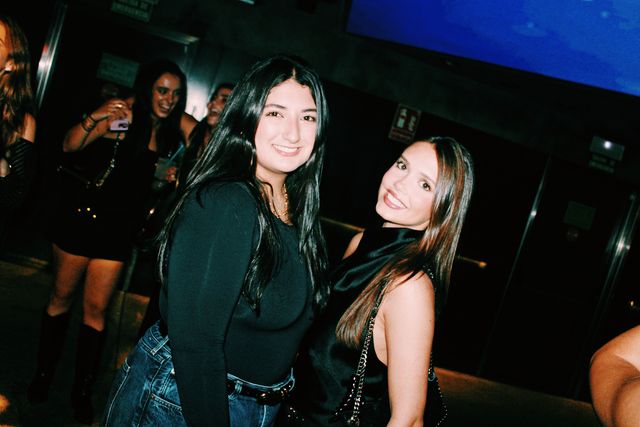 Dos chicas de negro posan juntas y sonríen a cámara, una con vaqueros y la otra mirando por encima del hombro, junto a unas puertas y bajo una pantalla azul.