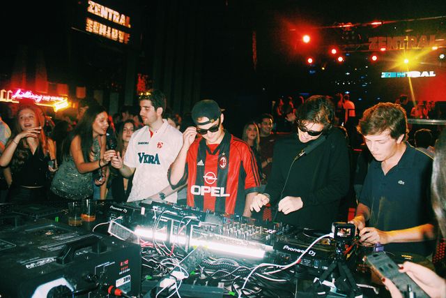 Tras la mesa de mezclas, el DJ de camiseta de fútbol a rayas rojas y negras se ajusta las gafas entre otros tres chicos, con focos rojos y letreros de Zentral detrás.