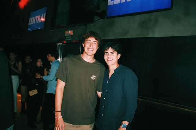 Dos chicos posan juntos y sonríen a cámara, uno con camiseta verde oliva y otro con camisa azul oscuro, bajo dos pantallas azules con el nombre de Zentral.
