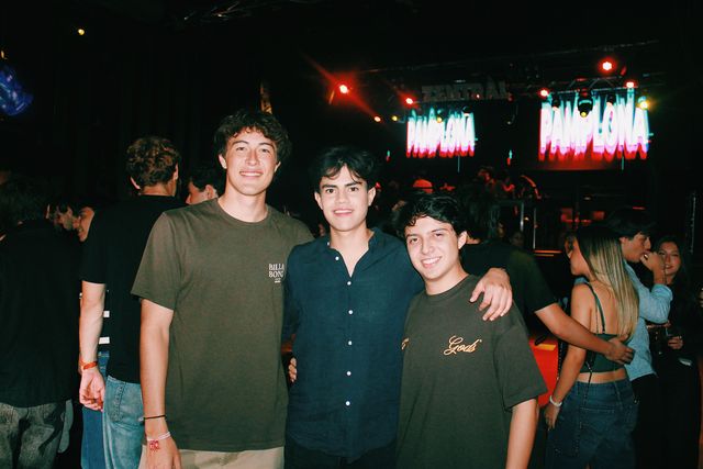 Tres chicos posan juntos y sonríen a cámara en la pista, con focos rojos y pantallas con la palabra Pamplona en letras blancas y rosas detrás.