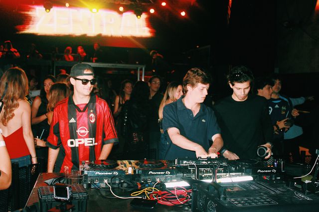 Tres chicos tras los platos bajo la pantalla de Zentral: el DJ de camiseta de fútbol a rayas rojas y negras sonríe, otro de polo azul mezcla y otro sujeta un micrófono.