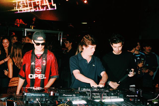 Un chico de polo azul mezcla entre el DJ de camiseta de fútbol a rayas rojas y negras, que sonríe, y un chico de negro con un micrófono en la mano.