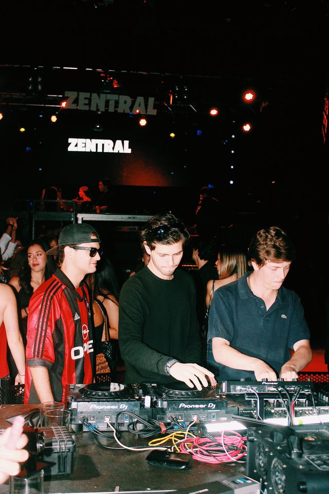 Dos chicos mezclan en los platos junto al DJ de camiseta de fútbol a rayas rojas y negras, de perfil, bajo dos letreros de Zentral y focos rojos.