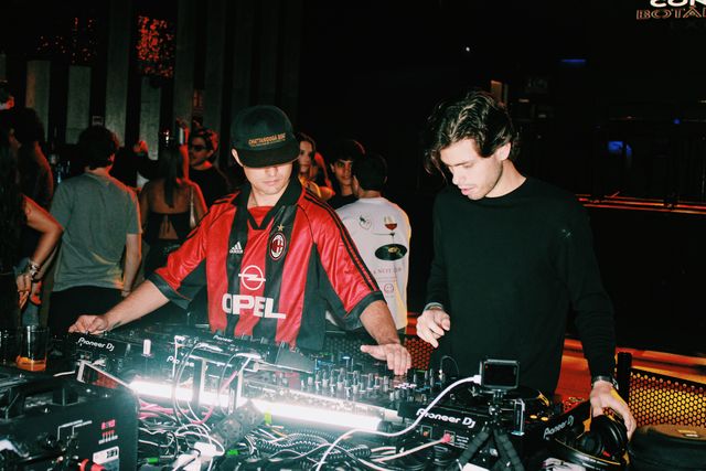 El DJ de camiseta de fútbol a rayas rojas y negras y un chico de jersey negro mezclan mirando los controles, con vasos sobre la cabina y gente detrás.