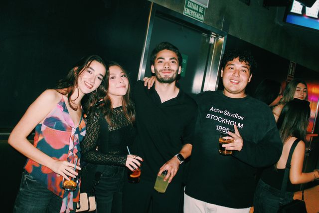 Dos chicas y dos chicos posan juntos con sus vasos y miran a cámara junto a una puerta de salida de emergencia; una chica lleva un top estampado de colores.