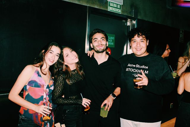 Dos chicas y dos chicos con vasos sonríen a cámara ante una salida de emergencia; la chica del top de encaje negro inclina la cabeza hacia la del top estampado.