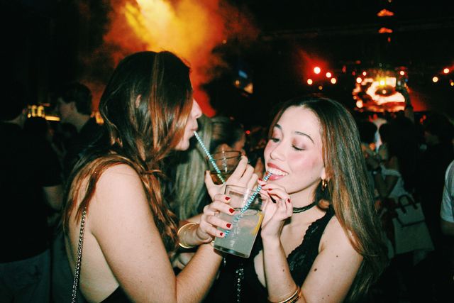 Dos chicas beben con pajitas de rayas azules y blancas, una de perfil y la otra sonriendo con los ojos entornados, entre humo naranja y luces rojas.