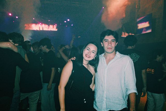 Una chica con top negro sonríe a cámara junto a un chico con camisa blanca que la rodea con el brazo, bajo el letrero de neón Zentral.