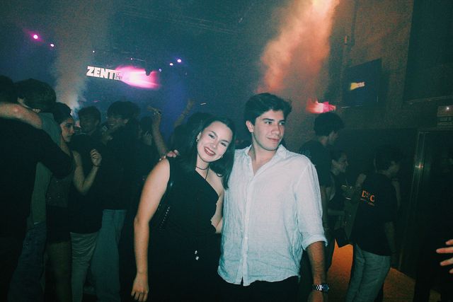 Una chica con top negro sonríe a cámara con la cabeza inclinada hacia un chico con camisa blanca que la abraza, con el público y una luz rosa detrás.