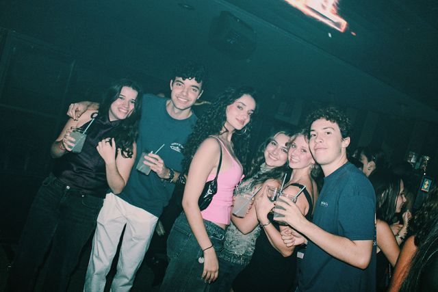 Cuatro chicas y dos chicos posan juntos y miran a cámara, la mayoría con un vaso con pajita en la mano, en una sala oscura de luz verdosa.