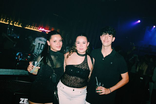 Dos chicas, una con top de rejilla y vaqueros blancos, y un chico con polo negro posan junto a la cabina del DJ, con focos azules al fondo.