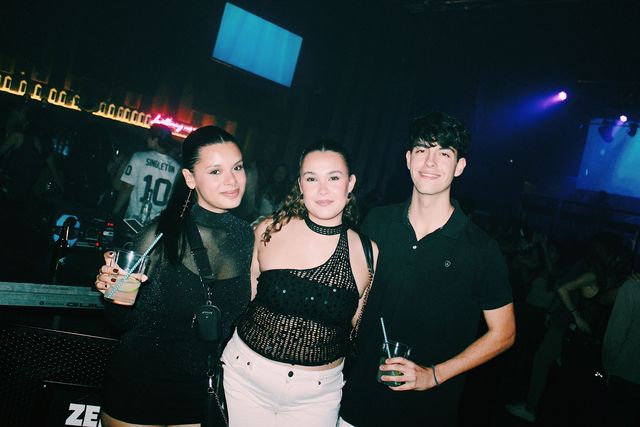 Dos chicas, una con top de rejilla y vaqueros blancos, y un chico con polo negro miran a cámara junto a la cabina del DJ, bajo una pantalla azul encendida.