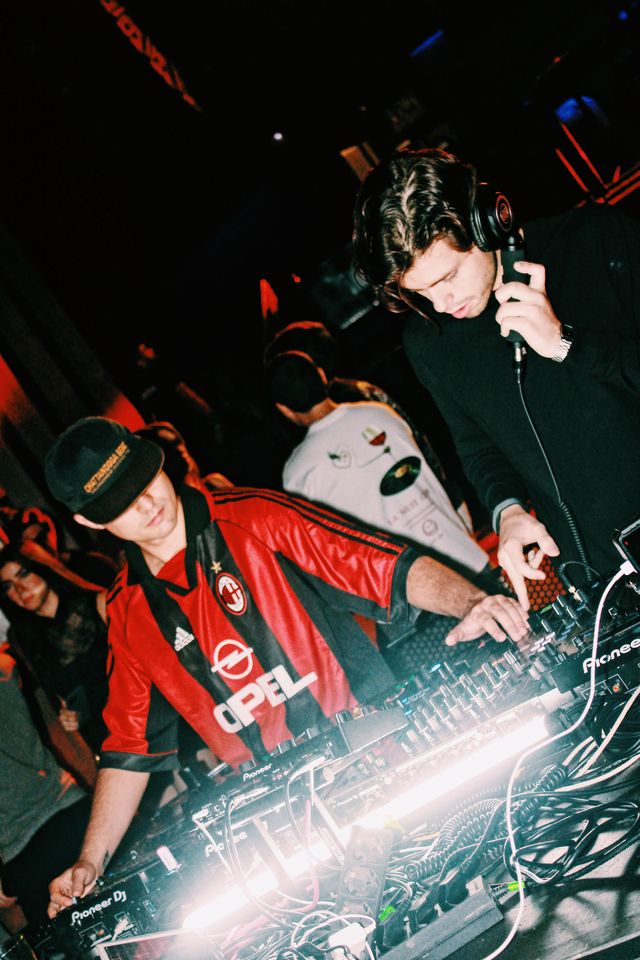 Con luces rojas al fondo, el DJ de camiseta de fútbol a rayas rojas y negras mezcla y, detrás, un chico de jersey negro escucha con un auricular de mano.