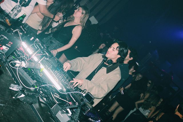 Un DJ con chaqueta beige y auriculares al cuello mezcla con la mirada baja junto a un tubo de luz blanca, con el público pegado a la cabina.