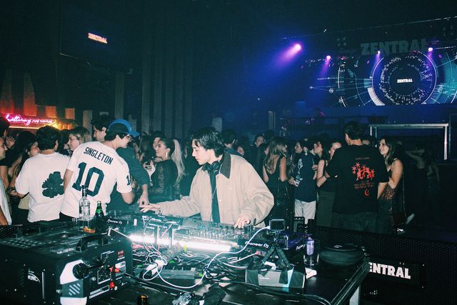 Un DJ con chaqueta beige mezcla en la cabina rodeado por el público, con una gran pantalla LED con el nombre de Zentral y focos violetas al fondo.