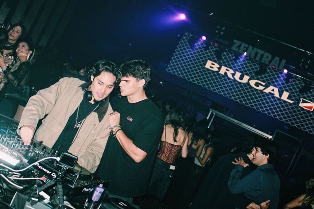 Un DJ con chaqueta beige sonríe en los platos mientras un chico con camiseta negra se inclina hacia él con los labios fruncidos, con una pantalla de Zentral al fondo.