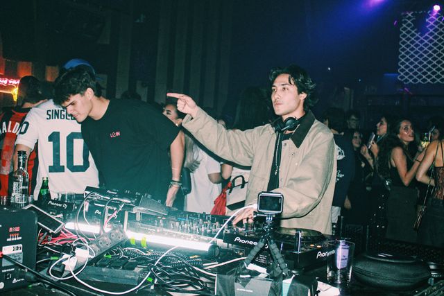 Un DJ con chaqueta beige señala con el dedo mientras un chico con camiseta negra se inclina sobre los platos, con el público alrededor.