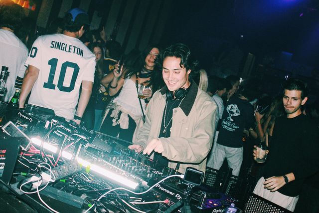 Un DJ con chaqueta beige sonríe mientras mezcla con las dos manos, rodeado de público; a la derecha, un chico de negro con un vaso mira a cámara.