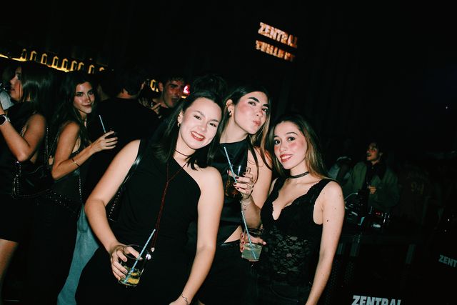 Tres chicas de negro posan juntas mirando a cámara; dos sonríen y la del centro pone morritos, con el letrero de Zentral al fondo.