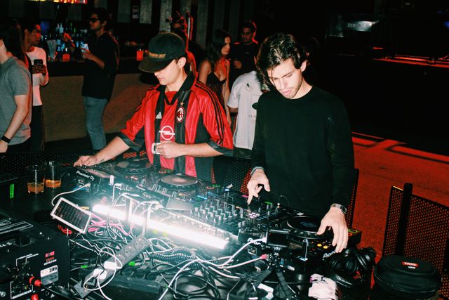 El DJ de camiseta de fútbol a rayas rojas y negras y un chico de jersey negro mezclan; al fondo, gente en la barra y un suelo iluminado en rojo.