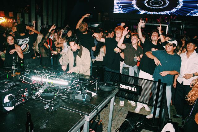 Un DJ con chaqueta beige mezcla con la cabeza baja mientras el público levanta los brazos a su alrededor, con una gran pantalla LED azul al fondo.