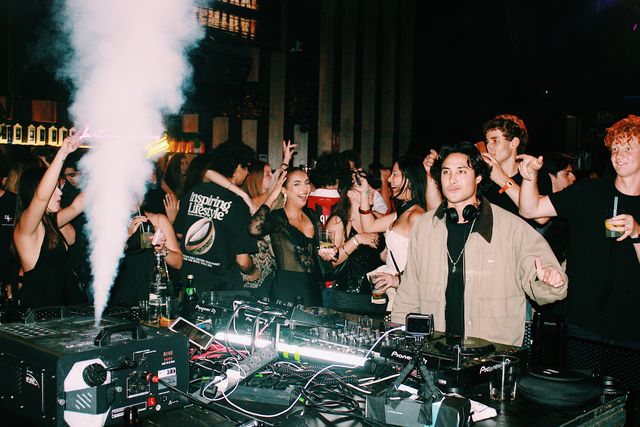 Un DJ con chaqueta beige mira a cámara y hace un gesto con la mano mientras una máquina lanza un chorro de humo blanco y el público levanta los brazos.