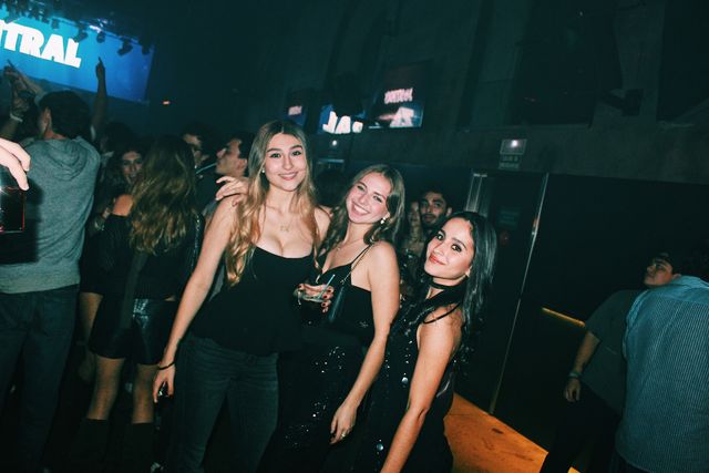 Tres chicas vestidas de negro posan juntas y sonríen a cámara, con el público a la izquierda y una puerta de emergencia y pantallas encendidas al fondo.