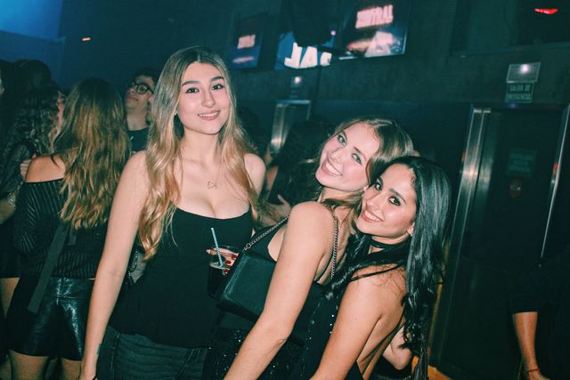 Tres chicas de negro sonríen a cámara; las dos de la derecha inclinan y juntan las cabezas, con pantallas encendidas y una puerta de emergencia detrás.