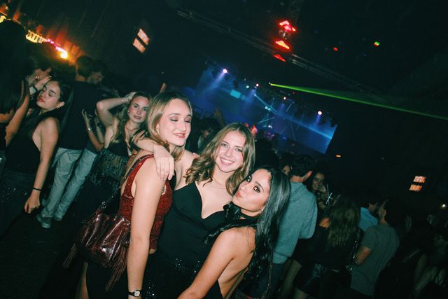 Tres chicas posan juntas: la de top granate cierra los ojos, la del centro sonríe a cámara y la de la derecha pone morritos, con un láser verde al fondo.