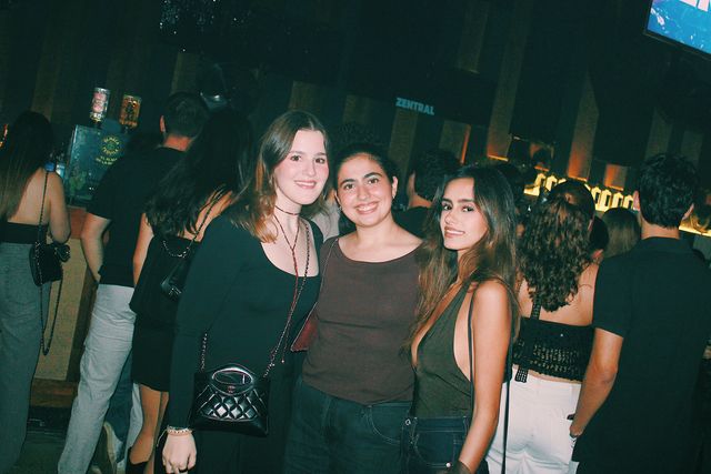 Tres chicas, una de negro, otra con camiseta marrón y otra con top verde oliva, sonríen a cámara con la barra iluminada en tonos cálidos al fondo.