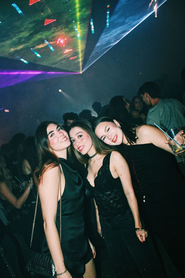 Tres chicas de negro juntan las cabezas y sonríen a cámara bajo un abanico de láseres verdes, azules y violetas, con el público entre el humo detrás.