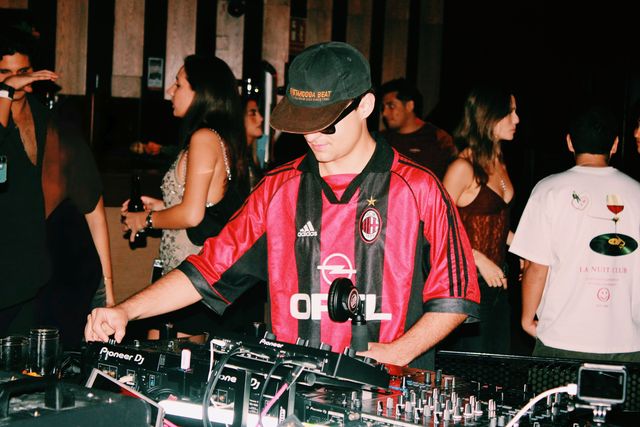 El DJ de camiseta de fútbol a rayas rojas y negras, con gorra y gafas de sol, mezcla con la cabeza baja mientras detrás charlan chicos y chicas.