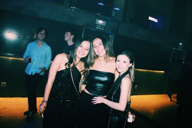 Tres chicas de negro, la del centro con top de cuero, posan juntas y sonríen a cámara; detrás, un chico con camisa azul claro sostiene un vaso.