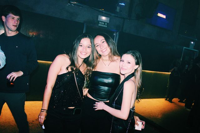 Tres chicas de negro, la del centro con top de cuero, sonríen a cámara muy juntas; a la izquierda, un chico con sudadera oscura sostiene un vaso.