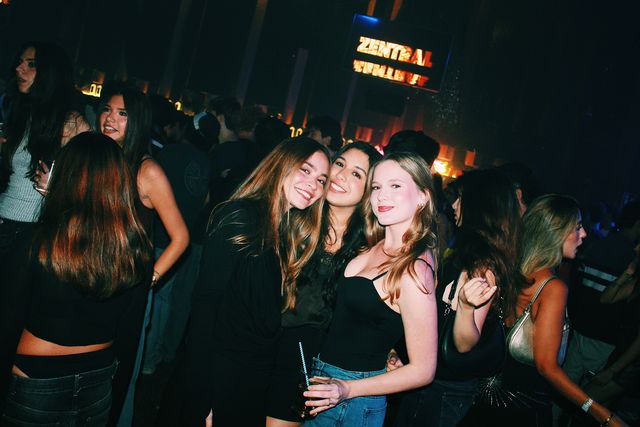 Tres chicas posan muy juntas y sonríen a cámara entre el público, una con un vaso con pajita, con el letrero de neón Zentral al fondo.