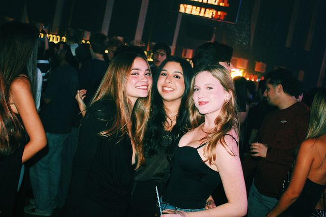 Tres chicas vestidas de negro posan juntas y sonríen a cámara entre el público, con una pantalla de letras naranjas al fondo; la de la derecha sostiene un vaso con pajita.