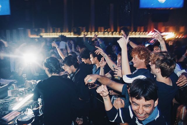 Un DJ de negro con manga larga mezcla de espaldas y a su lado otro chico con auriculares al cuello ríe, mientras el público levanta los brazos y señala hacia arriba.