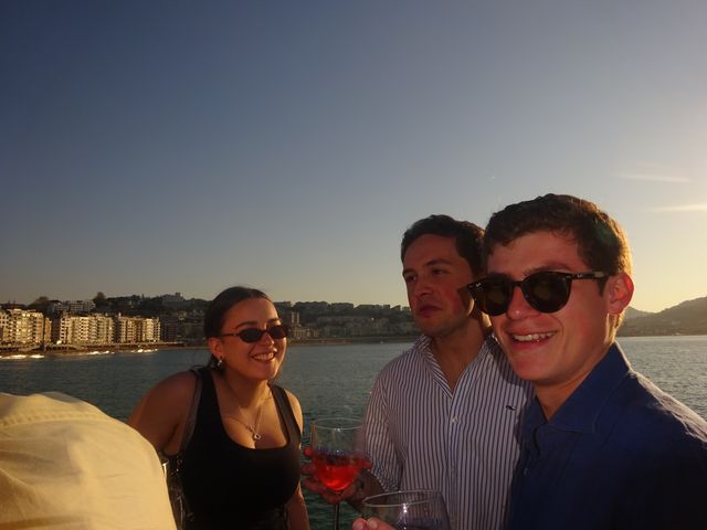 Una chica y dos chicos posan junto al mar al atardecer, con una copa de bebida roja en primer plano y la ciudad al fondo.