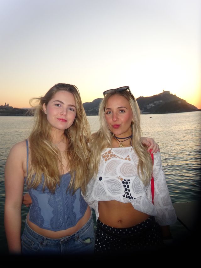 Dos chicas de pelo largo posan abrazadas junto al mar, con una colina recortada contra el cielo anaranjado del atardecer.