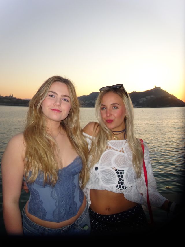 Dos chicas posan muy juntas frente al mar al atardecer, una con top azul de encaje y otra con blusa blanca y gafas de sol en el pelo.
