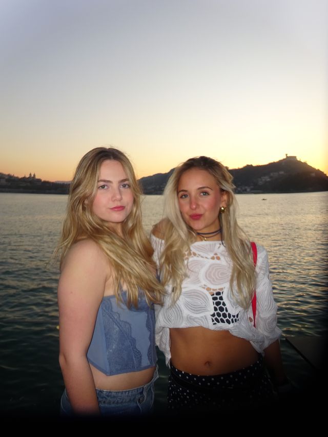 Dos chicas de pelo largo posan de pie junto al agua con la luz anaranjada del atardecer y una colina oscura al fondo.